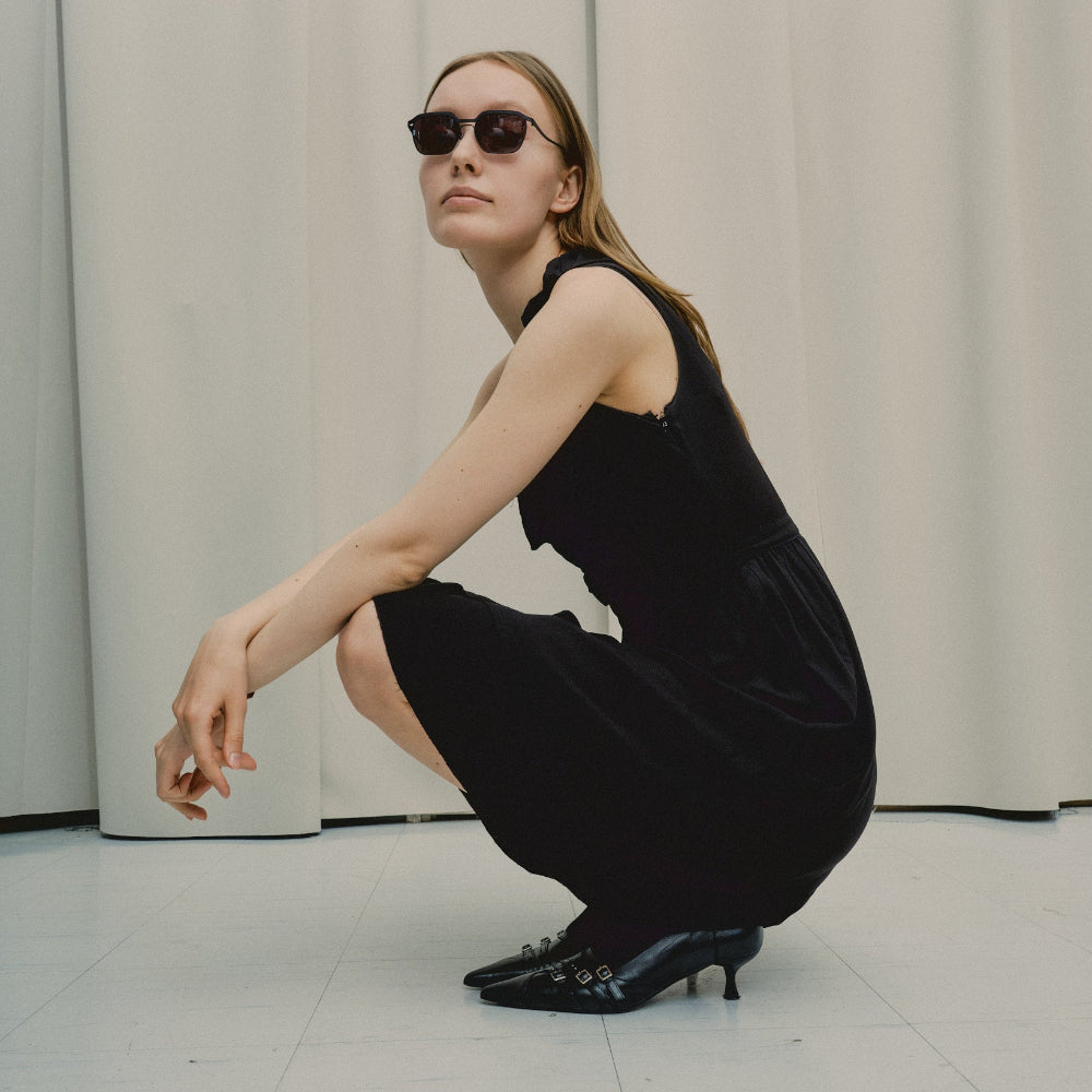 Model in a black dress crouching in the studio, wearing Vincent Catani Benno all-black sunglasses